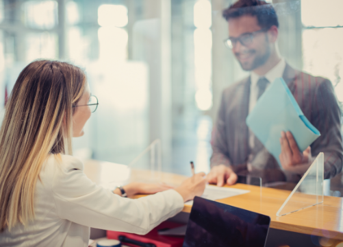 Bank teller assisting customer wearing glasses and holding a blue folder at the teller line.