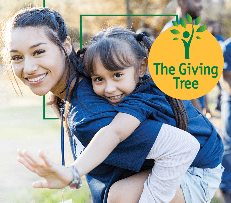 A woman outside with a braid in her hair giving a piggy back ride to a girl with pigtails and bangs who is waving toward the camera.