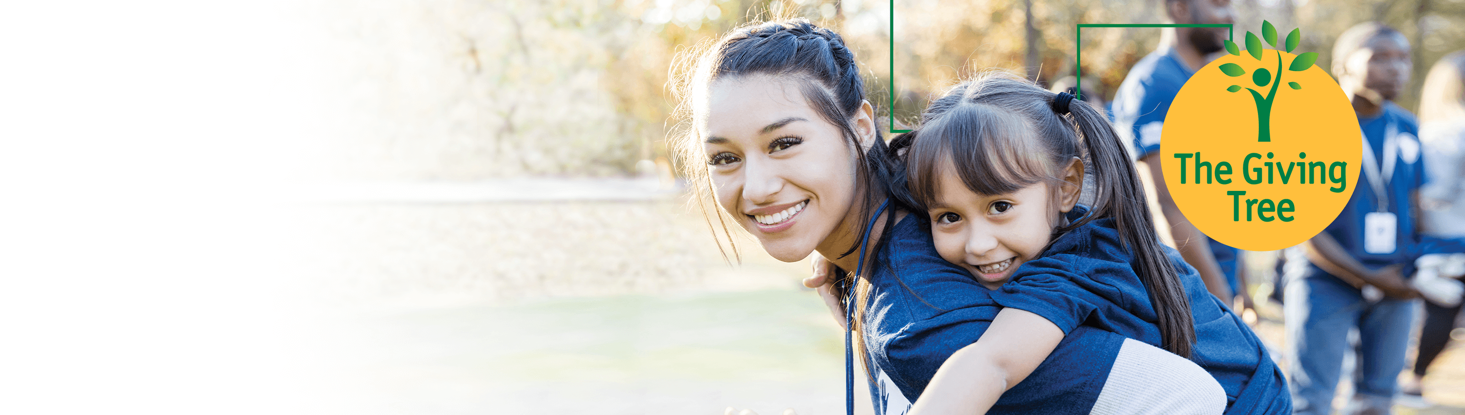 A woman outside with a braid in her hair giving a piggy back ride to a girl with pigtails and bangs who is waving toward the camera.