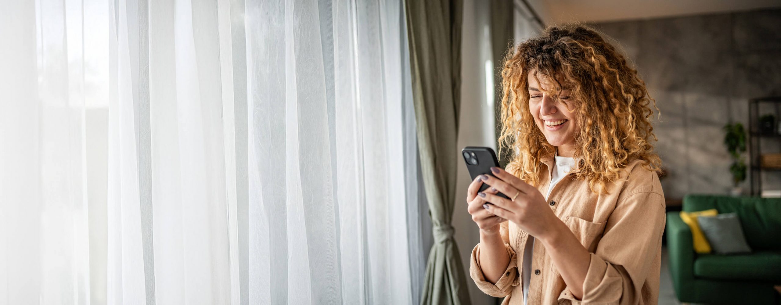 A women with curly hair standing near a window using her phone to open a checking account at TruNorth bank.
