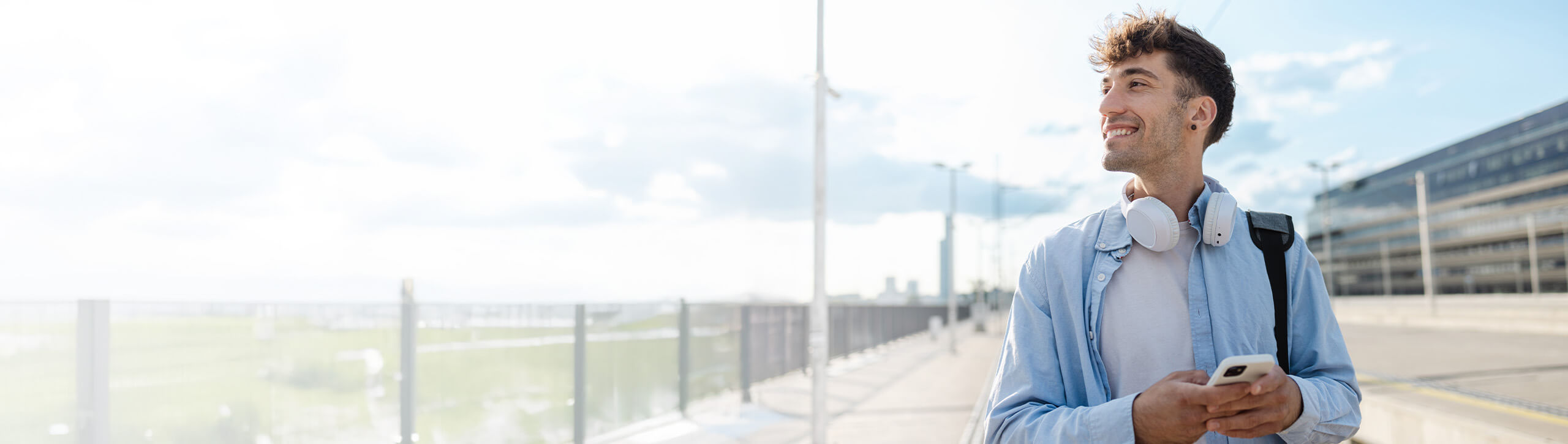 A man in a white t-shirt and blue dress shirt walking outside with headphones around his neck.