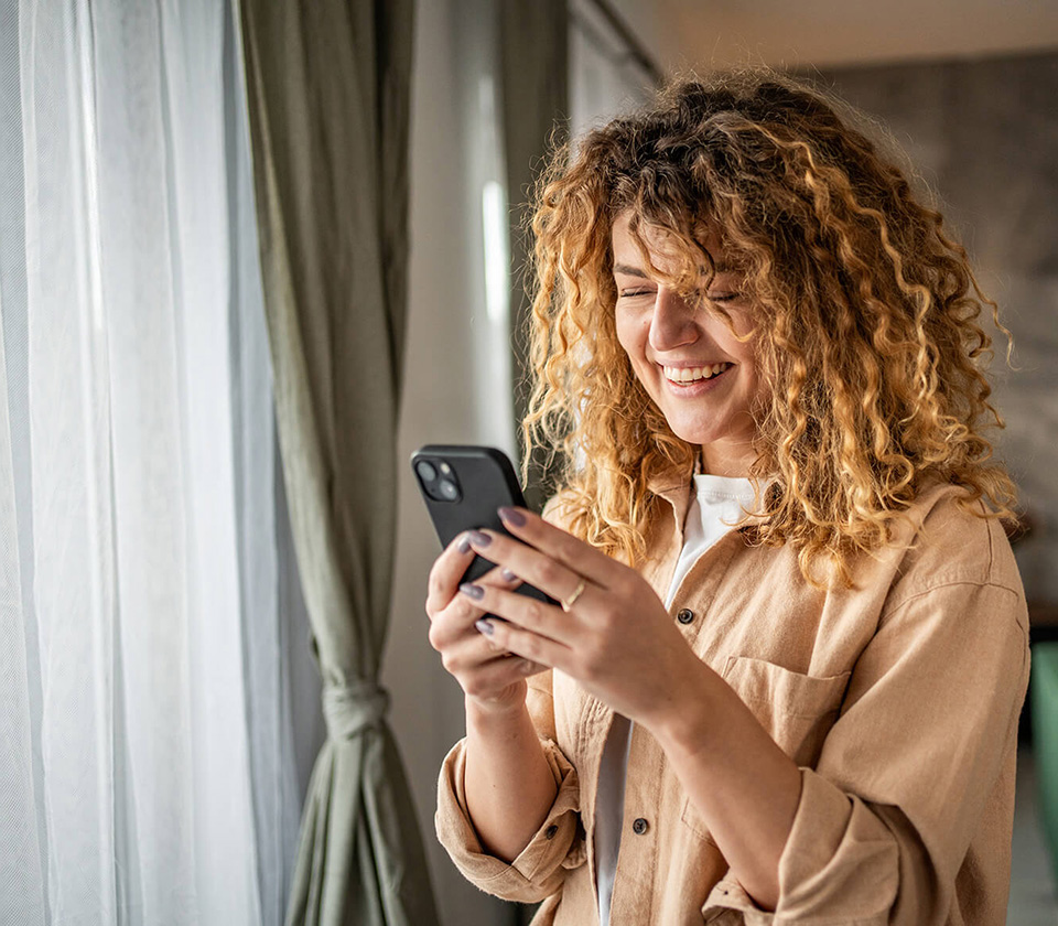 A woman with curly hair by a window looking at her phone